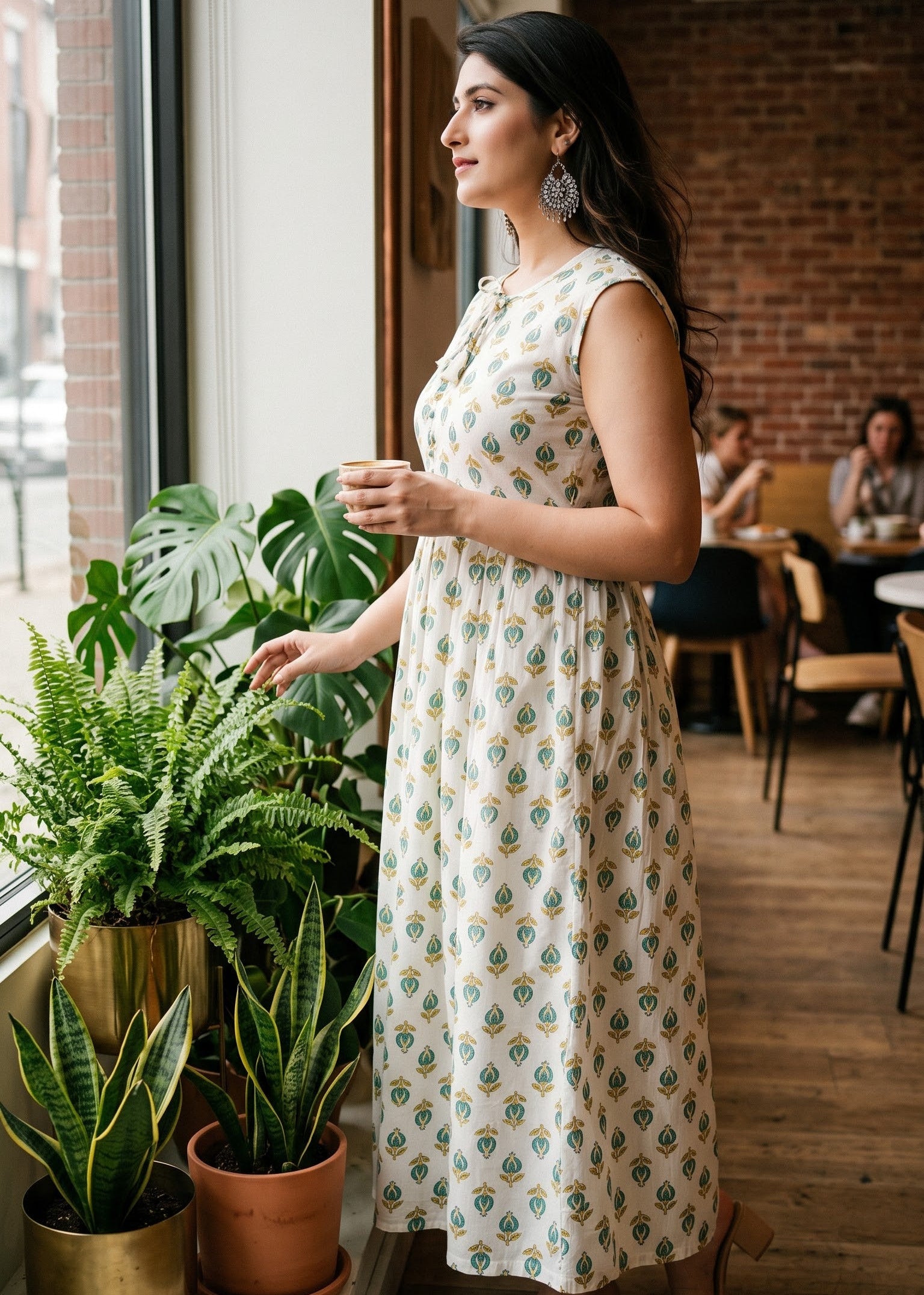 A woman wearing a sleeveless cream maxi dress with a green and blue geometric pattern stands by a window in a cafe, looking out while holding a small cup.
