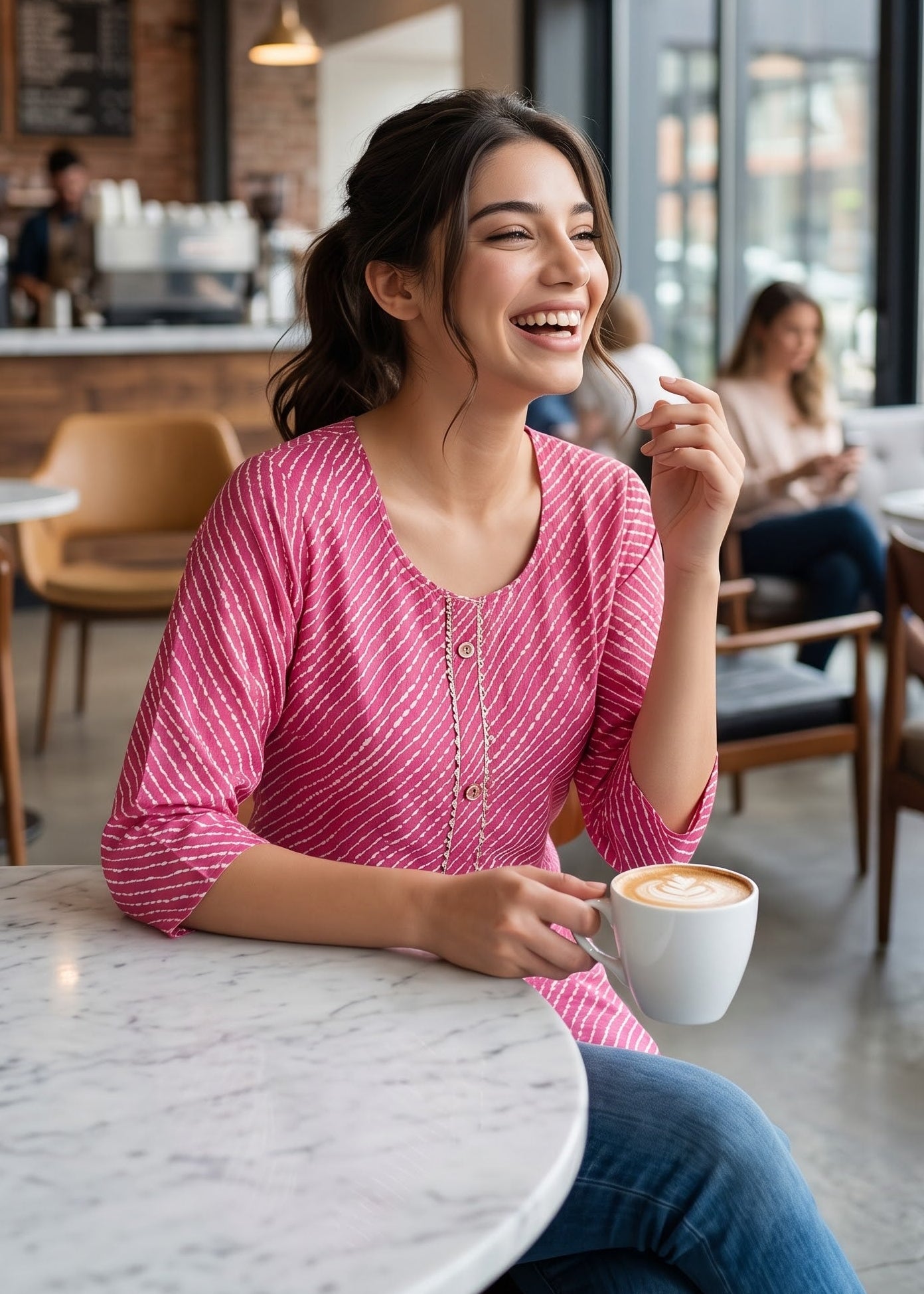A woman smiling and laughing while sitting at a white marble table in a modern coffee shop, wearing a pink and white lehriya patterned 3/4th sleeve top and blue jeans, holding a white coffee cup.