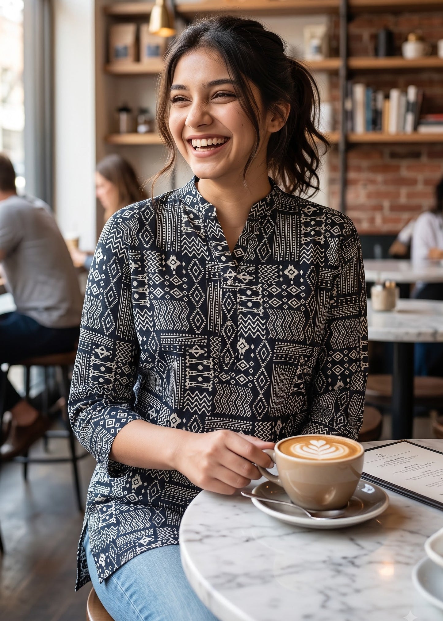 A woman smiling in a cozy cafe setting, wearing a black and white geometric patterned button-up short kurti tunic top and blue jeans, holding a cup of coffee with latte art.