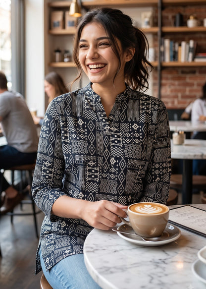 A woman smiling in a cozy cafe setting, wearing a black and white geometric patterned button-up short kurti tunic top and blue jeans, holding a cup of coffee with latte art.