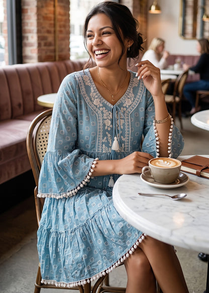 A woman laughing while sitting at a cafe table, wearing a light blue patterned short dress with white embroidery and pom-pom trim. She is holding a coffee cup on a marble-topped table in a cozy cafe setting with brick walls and warm lighting.