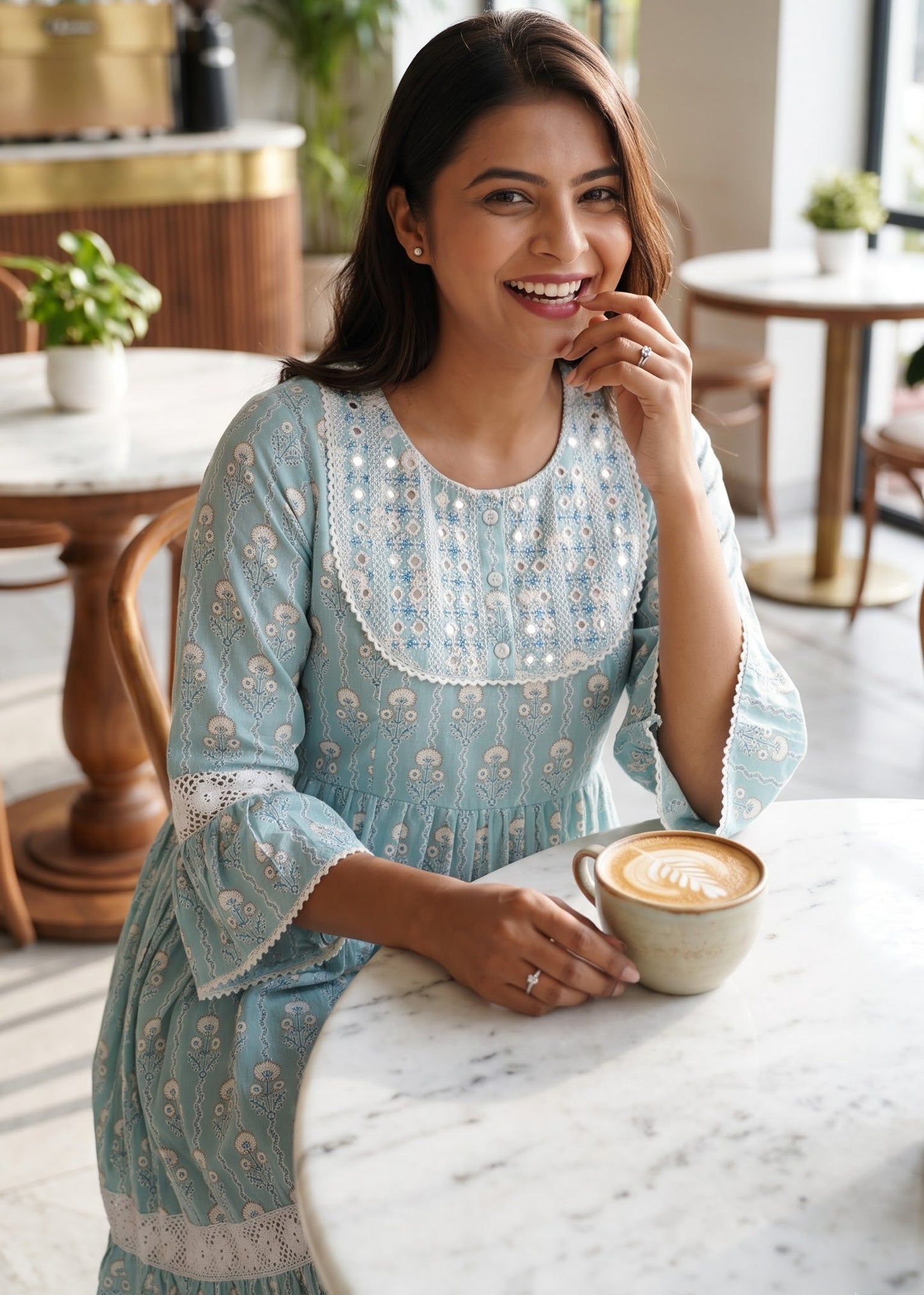 A woman wearing a light blue mirror embroidered short dress at a marble table in a cafe, holding a cup of coffee.
