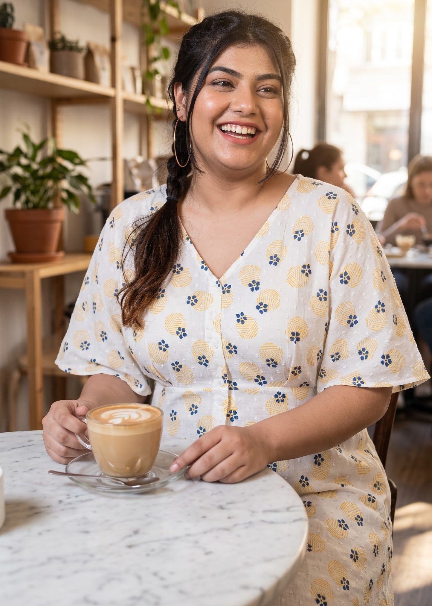 A woman smiling in a cozy cafe setting, wearing a white-colored V-neck dress with a yellow floral pattern and holding a cup of latte art on a marble table.