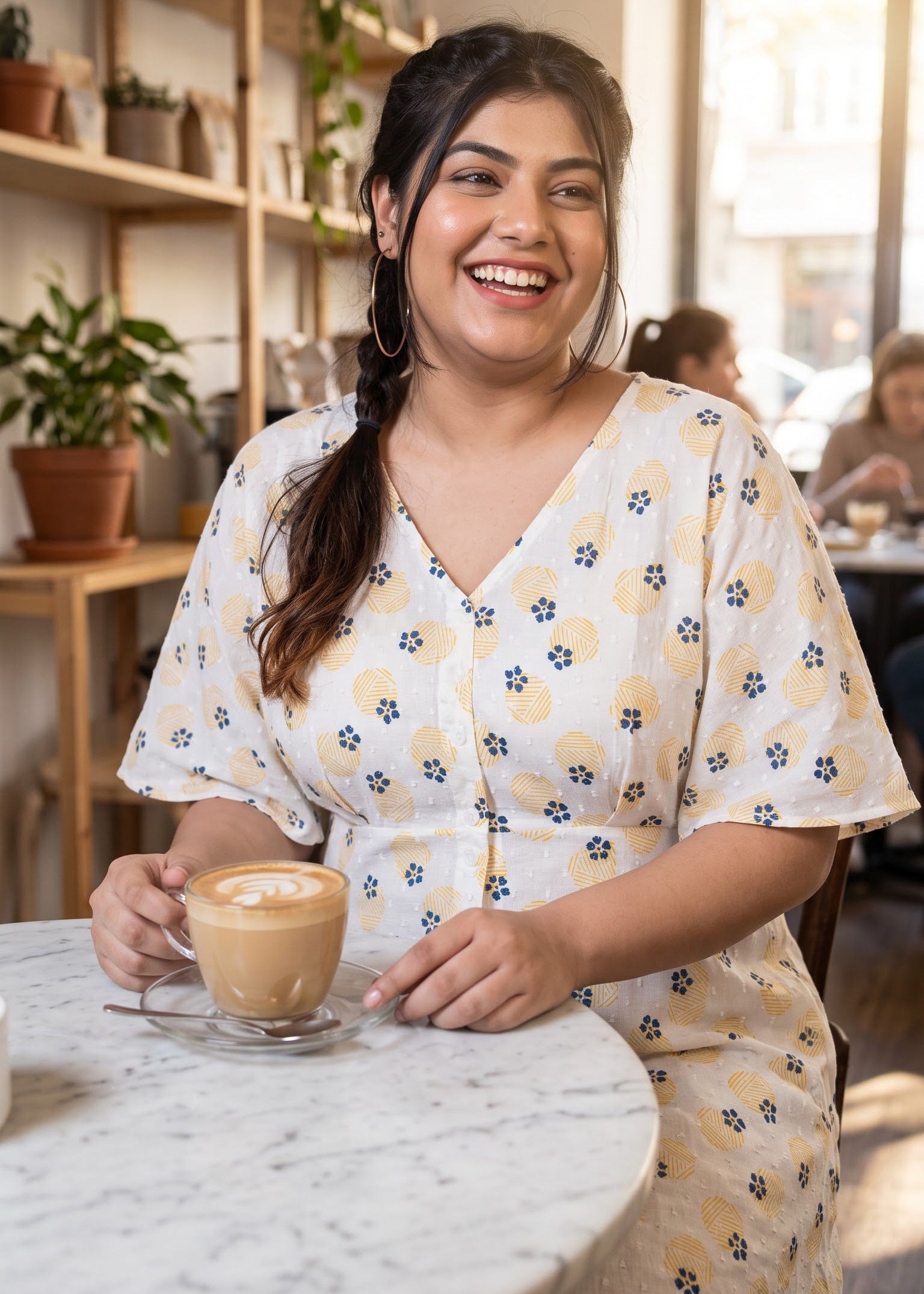 A woman smiling in a cozy cafe setting, wearing a white-colored V-neck dress with a yellow floral pattern and holding a cup of latte art on a marble table.