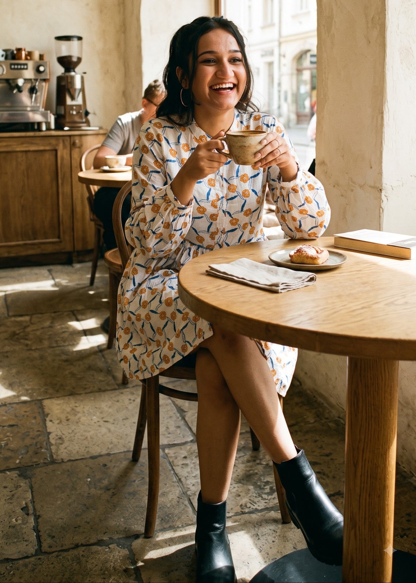 A woman smiling while sitting at a wooden table in a cafe, holding a cup. She is wearing a white-colored dress with a blue and orange abstract floral pattern, black ankle boots.