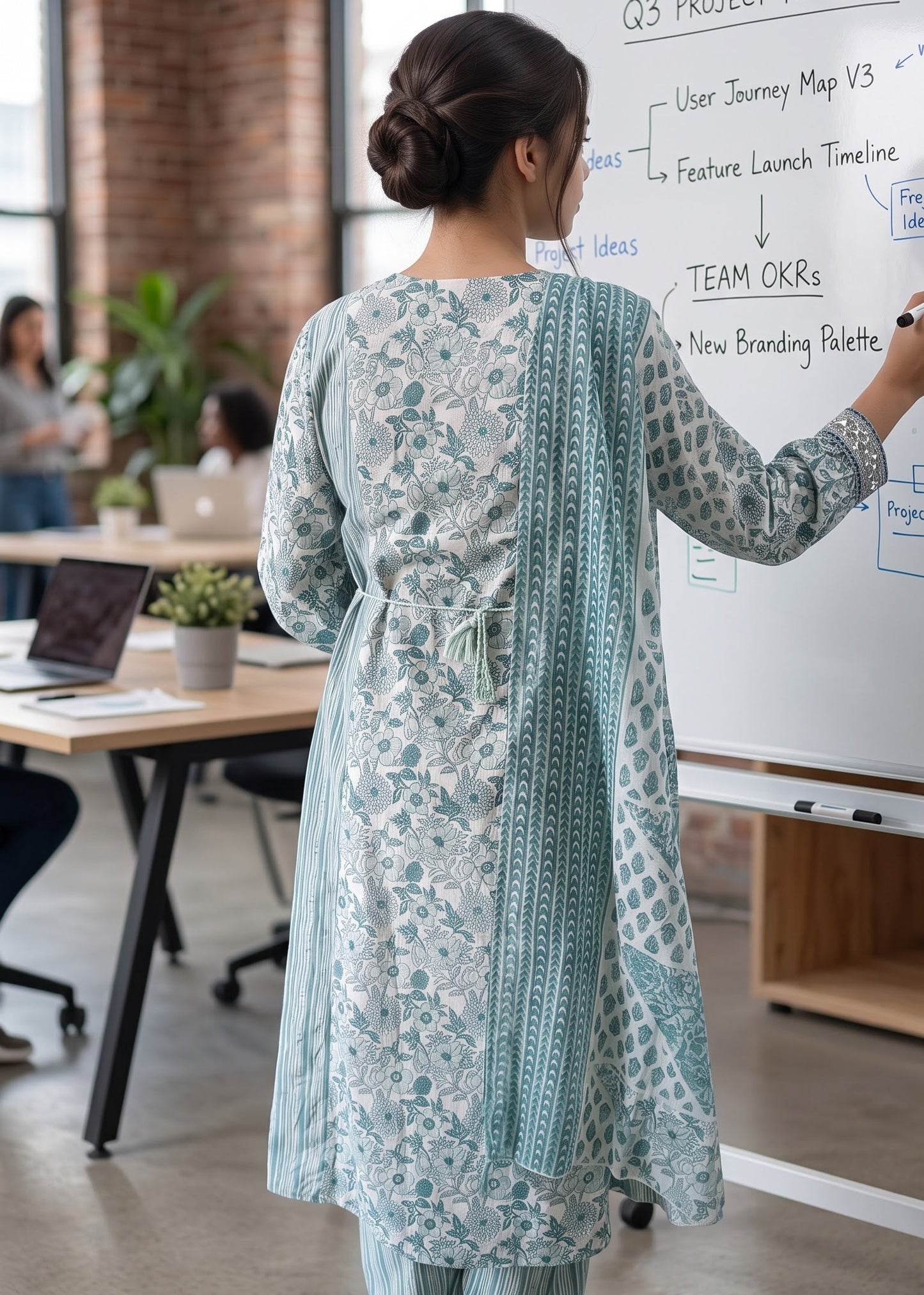 A woman stands with her back to the camera in a bright office setting, wearing a light blue and white floral printed kurta with back tie up, matching stripe trousers and a patterned dupatta.