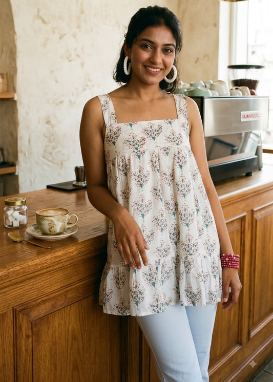 A woman wearing a sleeveless white tiered tunic top with a delicate blue and pink floral print, paired with light blue jeans, standing in a cozy cafe setting with wooden counters and hanging brass lamps.