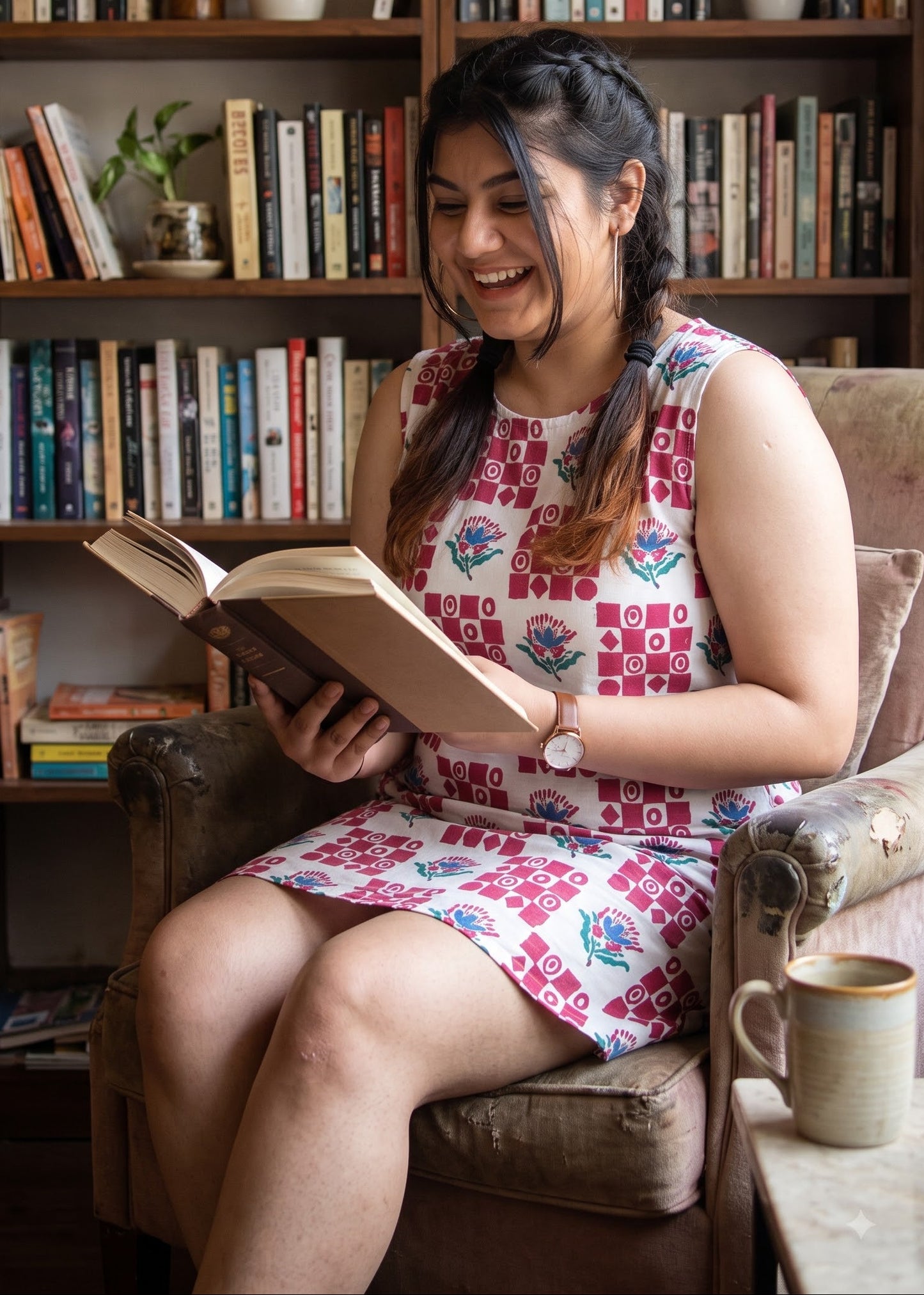 A woman with braided hair sits in a brown armchair, smiling while reading an open book in front of a bookshelf. She is wearing a sleeveless white dress with a red, white and blue geometric pattern.