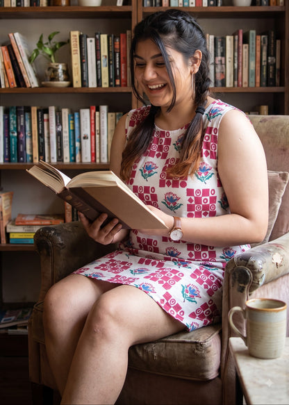 A woman with braided hair sits in a brown armchair, smiling while reading an open book in front of a bookshelf. She is wearing a sleeveless white dress with a red, white and blue geometric pattern.