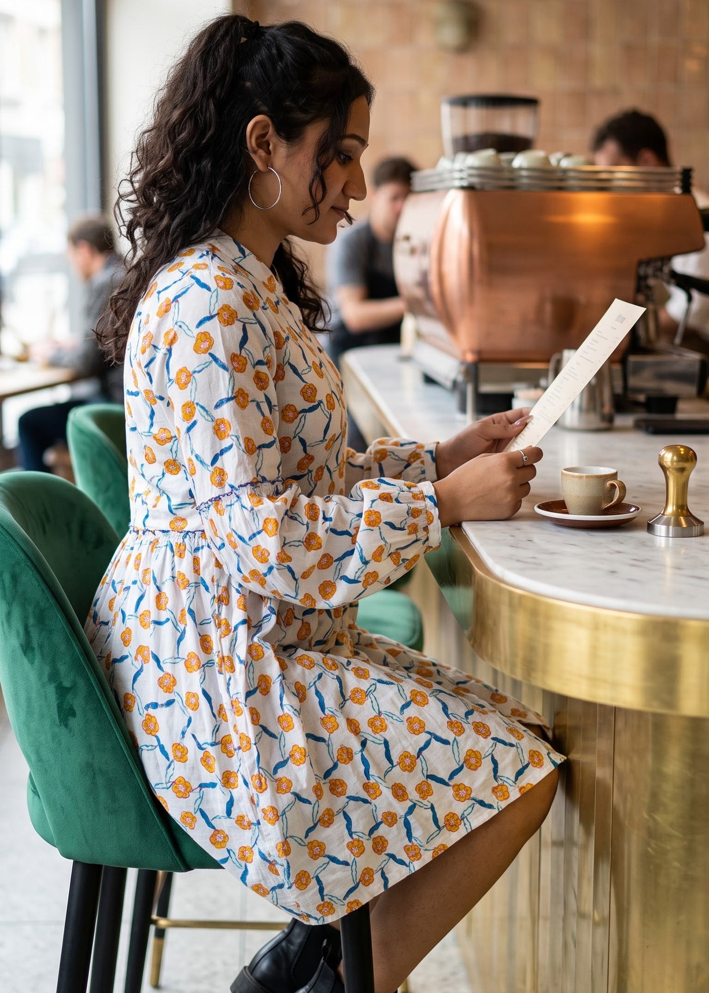 A woman sits at a cafe counter reading a menu, wearing a white long puff-sleeve dress with a yellow and blue floral pattern. She is seated on a green velvet bar stool in a warm, modern cafe setting with a copper coffee machine in the background.