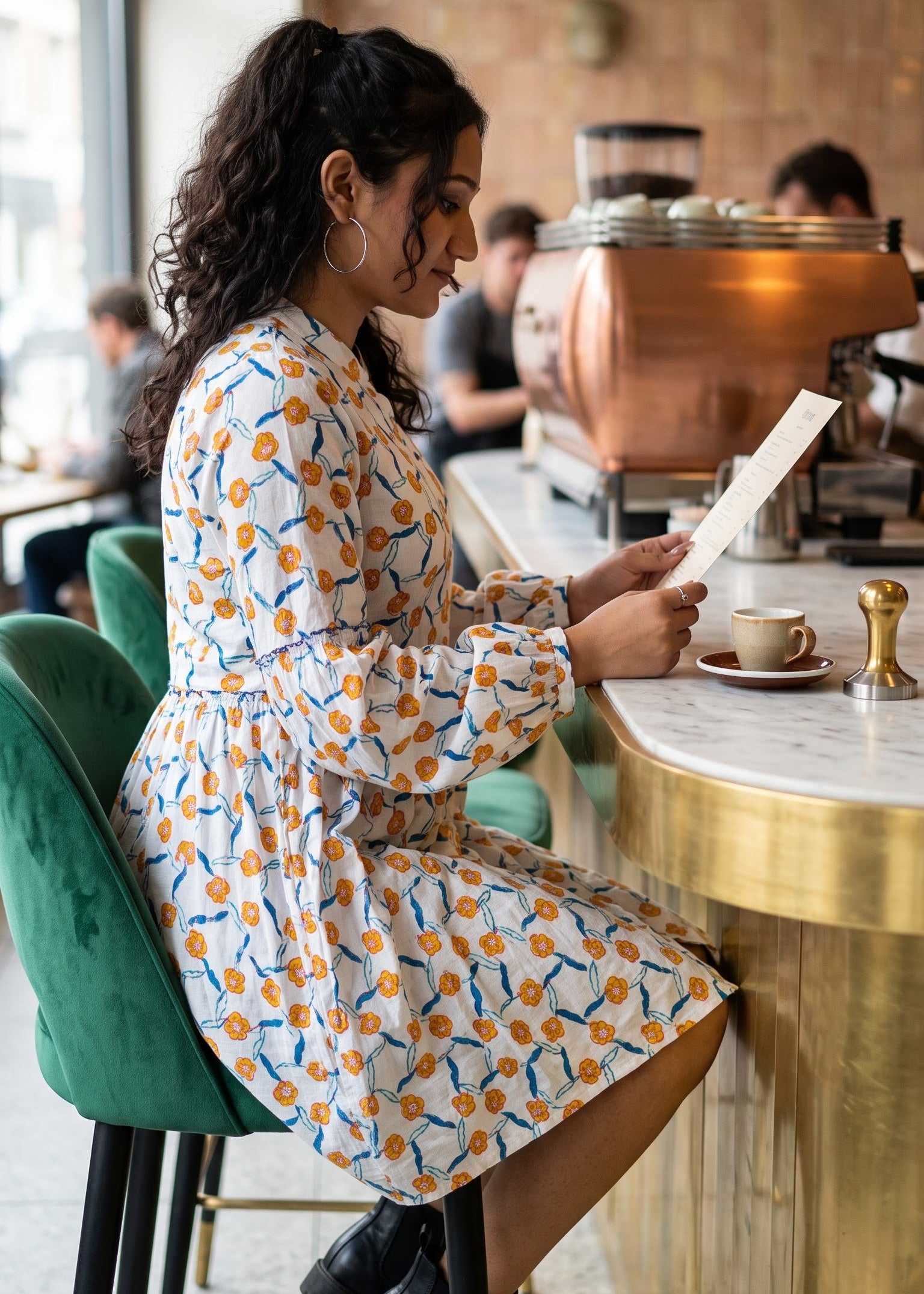 A woman sits at a cafe counter reading a menu, wearing a white long puff-sleeve dress with a yellow and blue floral pattern. She is seated on a green velvet bar stool in a warm, modern cafe setting with a copper coffee machine in the background.