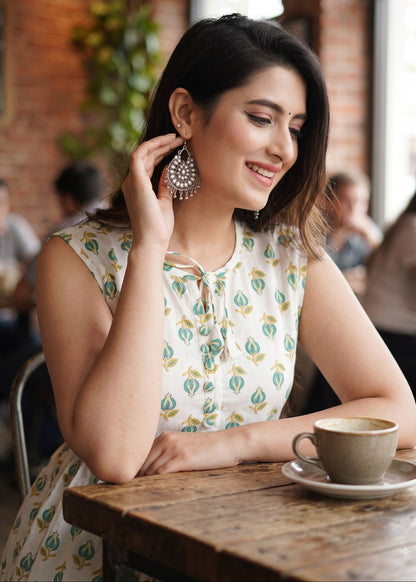 A woman in a cafe wearing a cream sleeveless maxi dress with a green and yellow floral print, holding a silver-toned statement earring near her ear. A coffee cup sits on the wooden table in front of her.