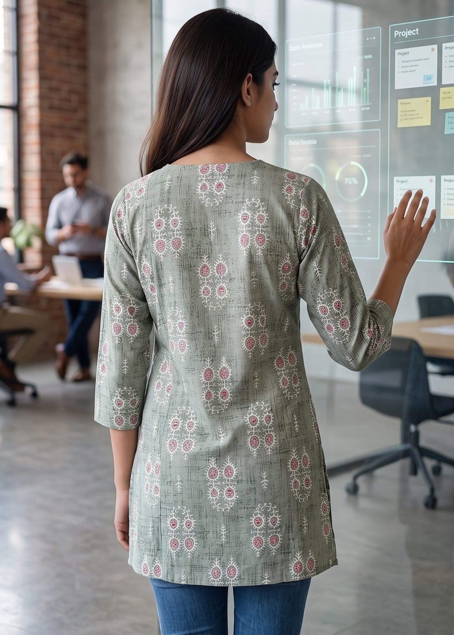 A woman viewed from behind wearing a sage green three-quarter sleeve tunic short kurti top with a red and white floral pattern, standing in a modern office and touching a glass display board.
