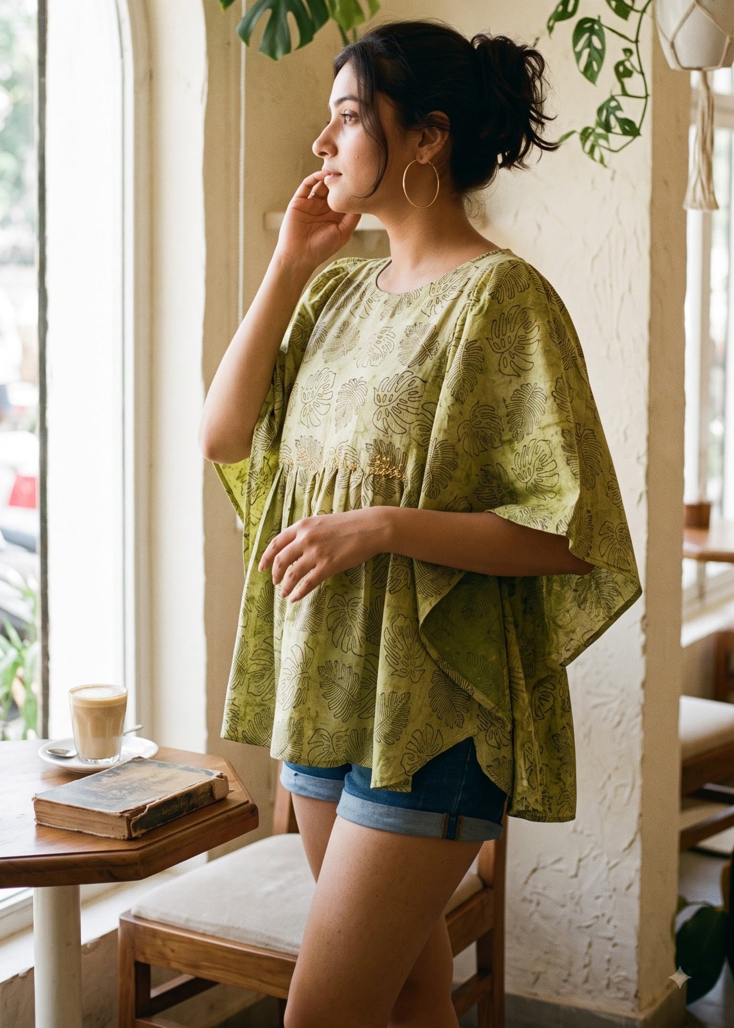 A woman wearing a loose, olive-green patterned tunic top and denim shorts stands by a window in a bright, modern cafe setting.