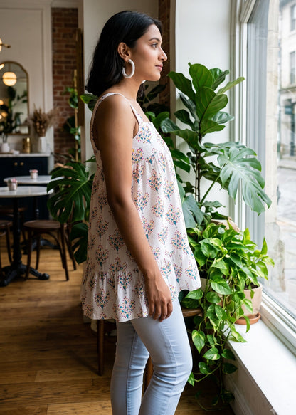 A woman wearing a sleeveless floral top and light blue jeans stands in profile by a large window in a bright indoor space with potted plants.