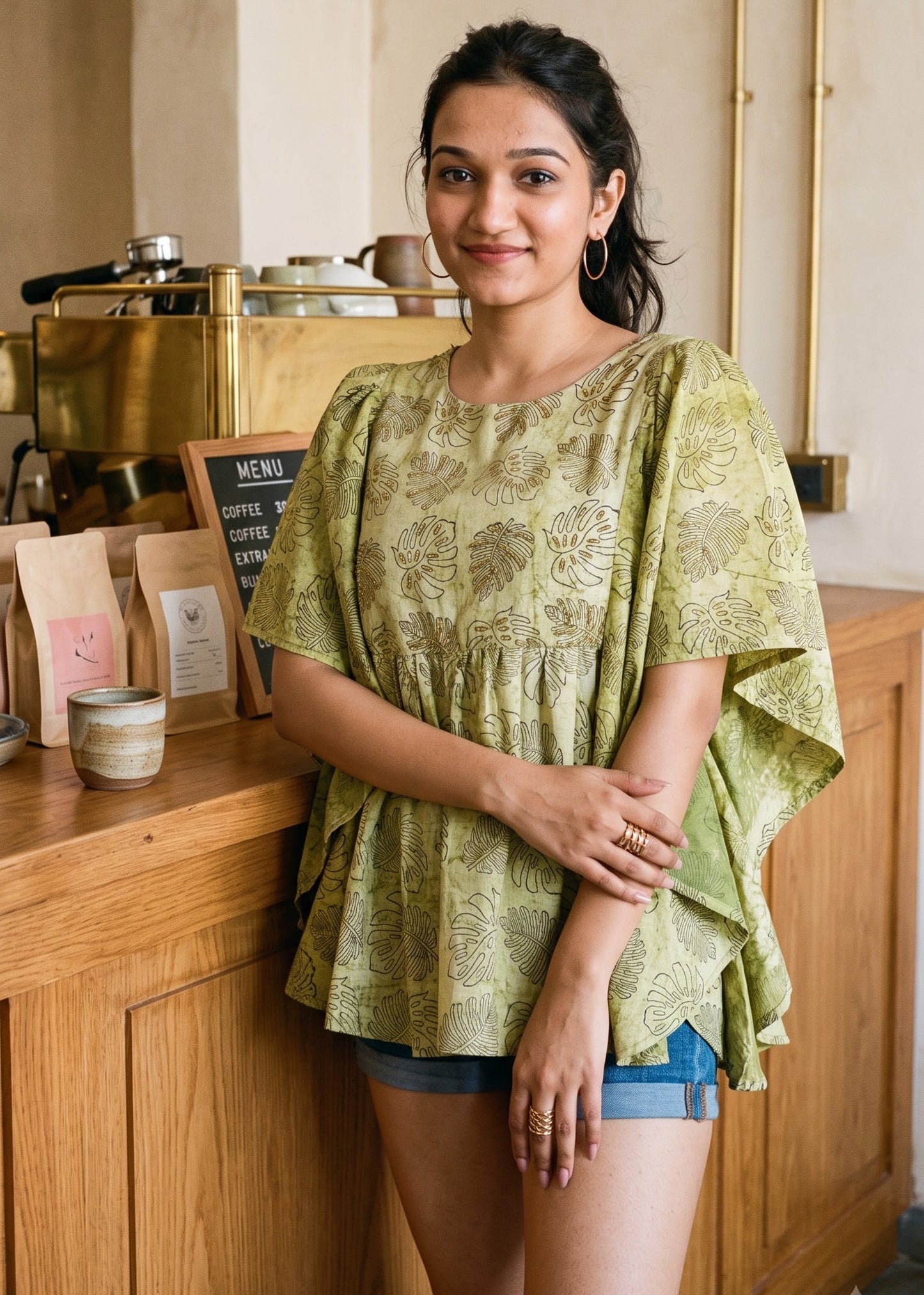 A woman wearing a light olive green patterned short butterfly-sleeve tunic top and denim shorts stands in a warm, rustic cafe setting with wooden counters and brass fixtures.