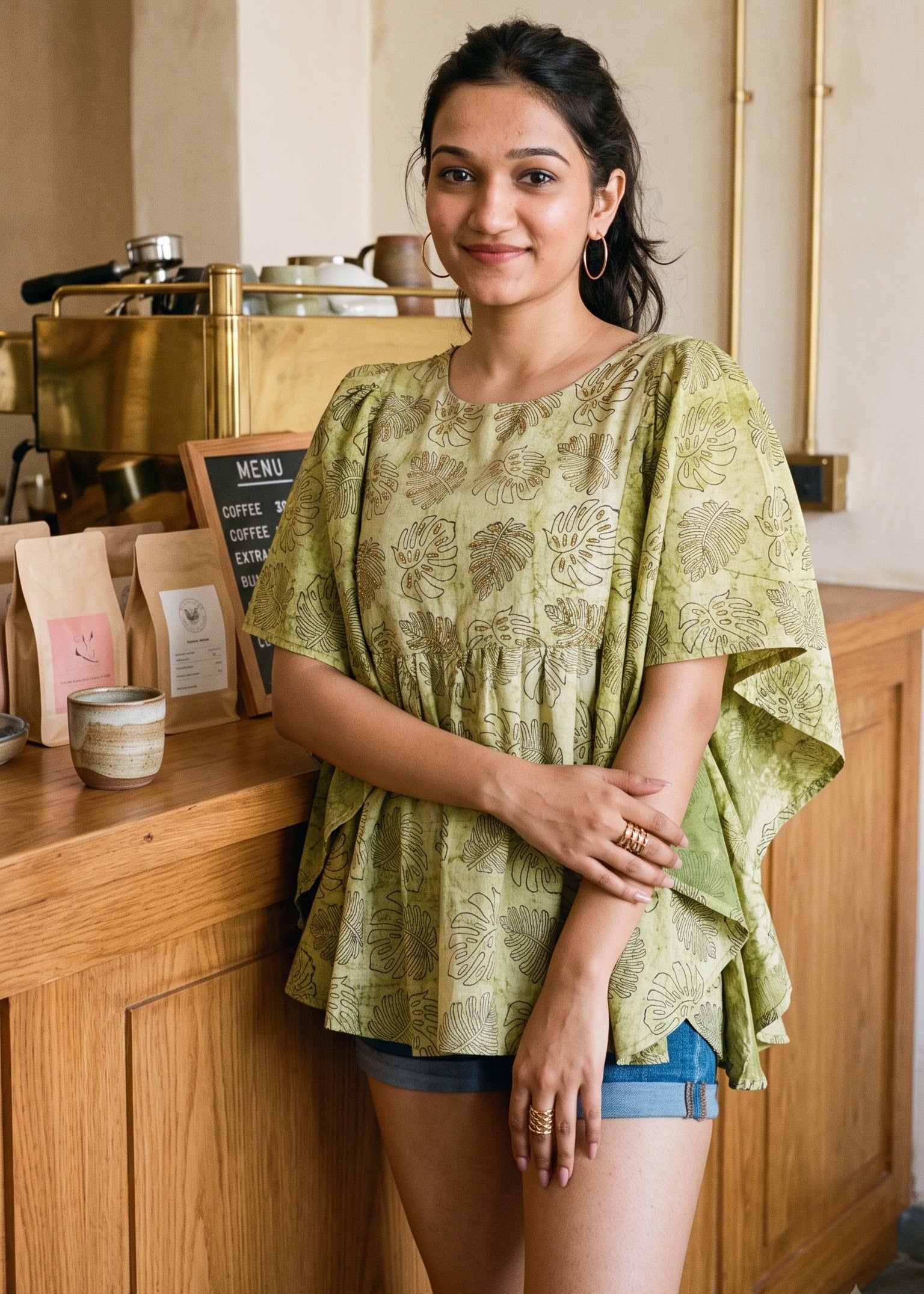 A woman wearing a light olive green patterned short butterfly-sleeve tunic top and denim shorts stands in a warm, rustic cafe setting with wooden counters and brass fixtures.