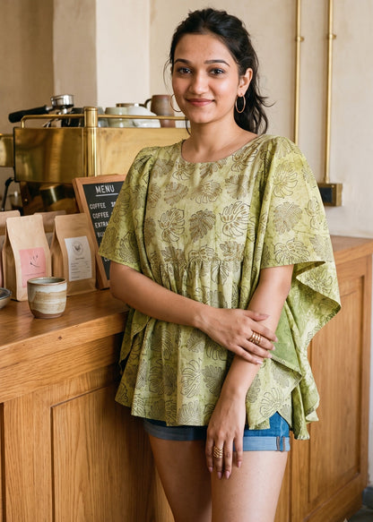 A woman wearing a light olive green patterned short butterfly-sleeve tunic top and denim shorts stands in a warm, rustic cafe setting with wooden counters and brass fixtures.