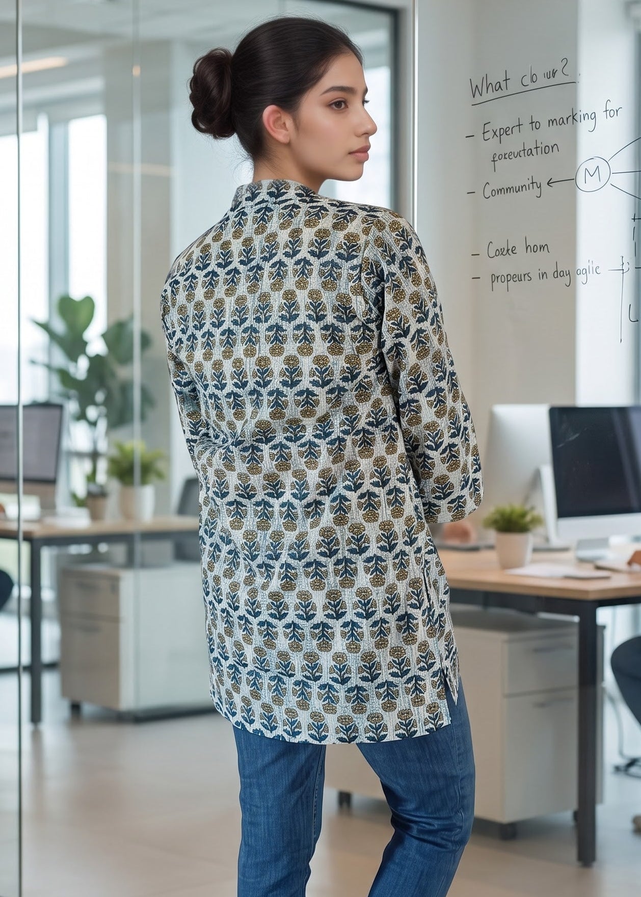 A woman stands in a modern office wearing a 3/4th-sleeve tunic short kurti top with a blue, beige, and white floral pattern, paired with blue jeans and white heels.
