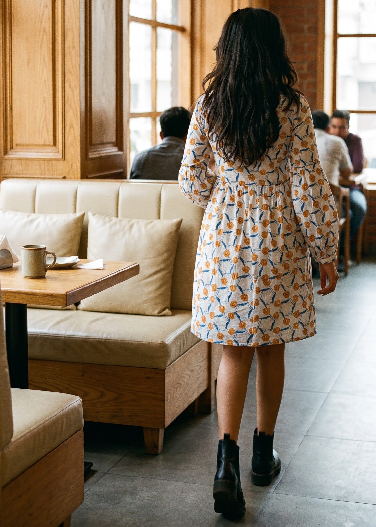A woman viewed from behind wearing a white-colored mini dress with a colorful geometric pattern, walking through a cozy cafe with wood paneling and brick walls.