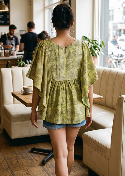 A woman viewed from behind wearing a loose, short butterfly-sleeve green patterned kaftan top and denim shorts while walking through a bright cafe with cream-colored booths and wooden floors.