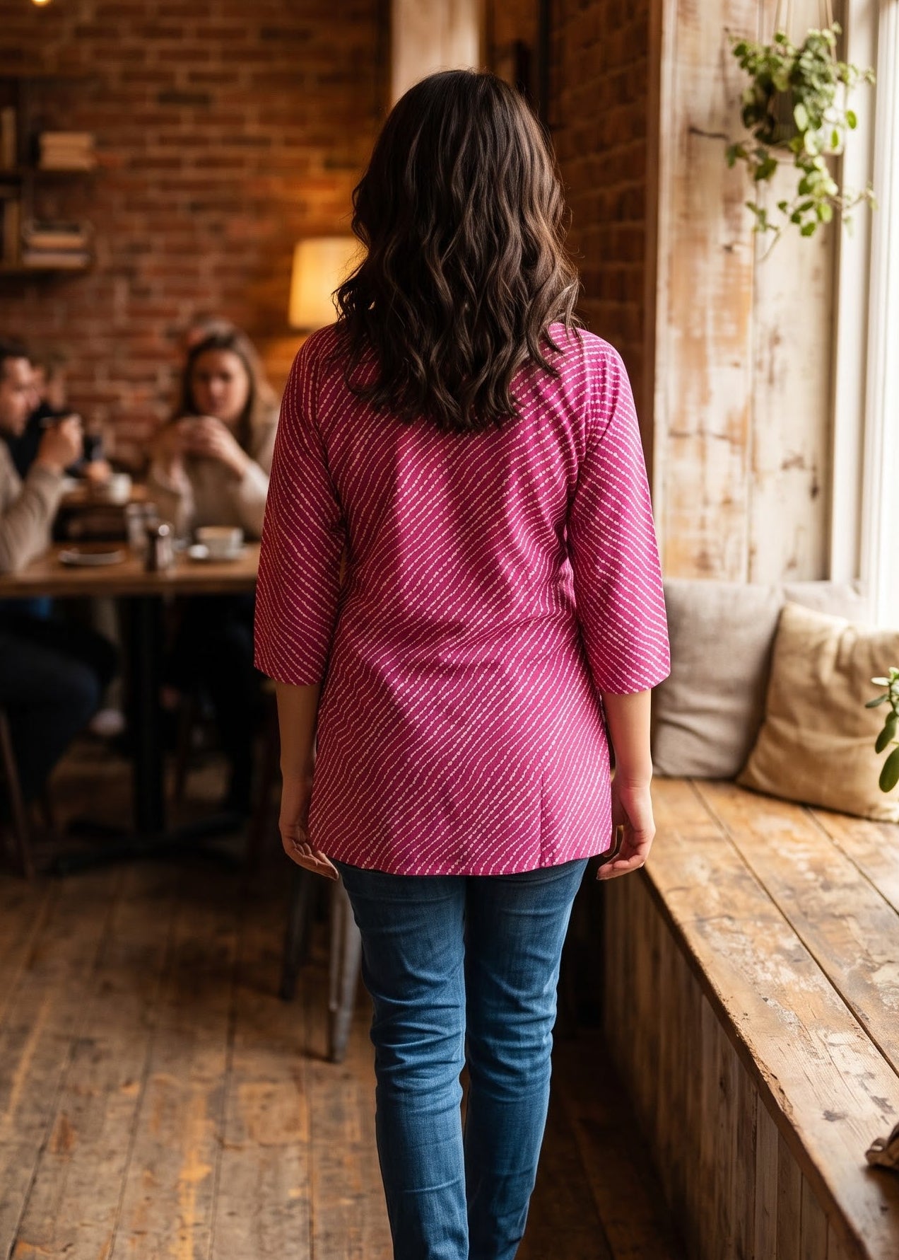A woman viewed from behind walking down a rustic wooden floor in a cozy cafe, wearing a pink and white lehriya patterned top and blue jeans.