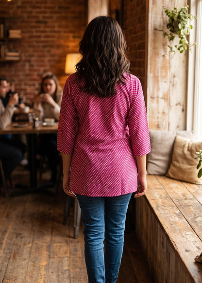 A woman viewed from behind walking down a rustic wooden floor in a cozy cafe, wearing a pink and white lehriya patterned top and blue jeans.