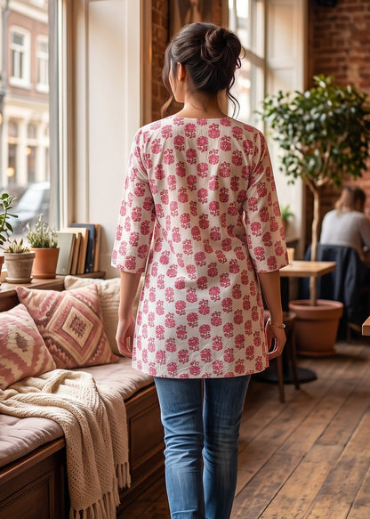 A woman viewed from behind wearing a pink and white patterned short kurti tunic top with blue jeans, walking down a wooden floor in a cozy, sunlit cafe with brick walls and warm hanging lights.