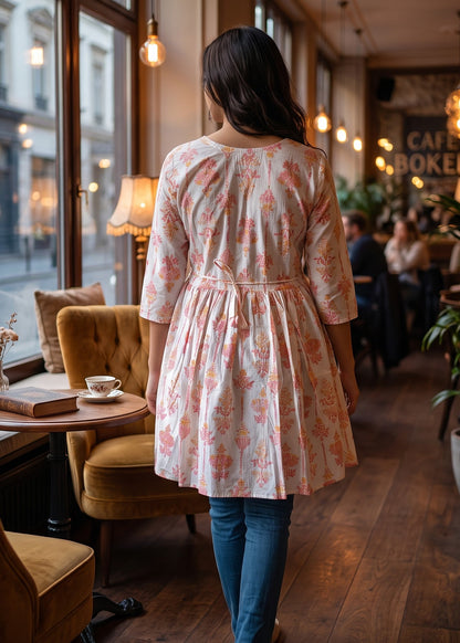 A woman viewed from behind wearing a light orange and white floral print tunic dress with three-quarter sleeves, back tie-up, standing in a cozy, warmly lit cafe with wooden floors and yellow velvet chairs.