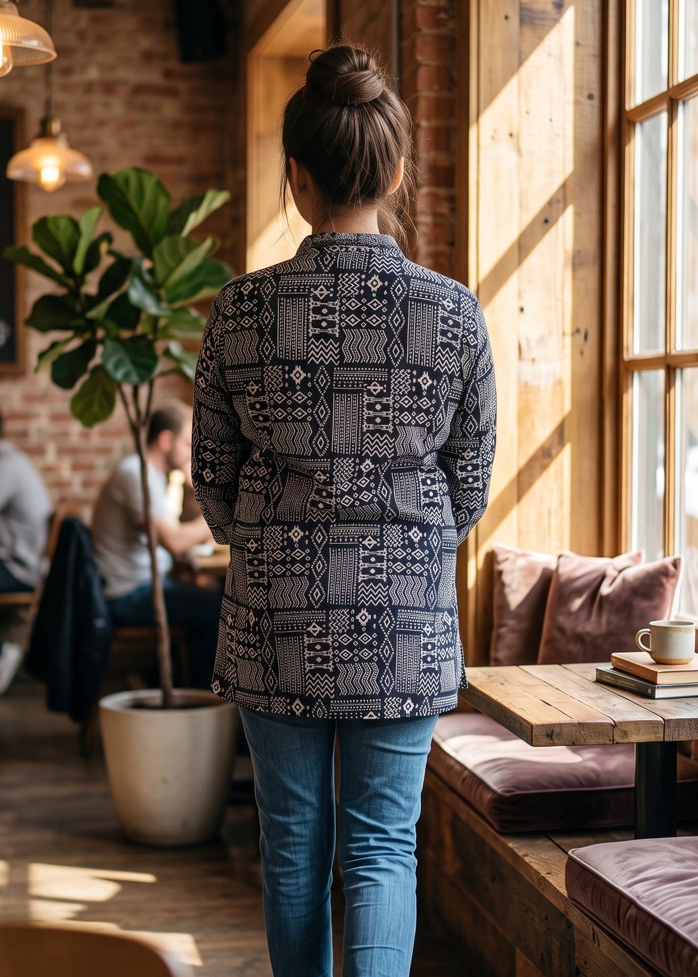 A person viewed from behind wearing a black and white geometric patterned button-up short kurti tunic top, blue jeans, and white sneakers, walking inside a rustic cafe with wood floors, brick walls, and large windows.