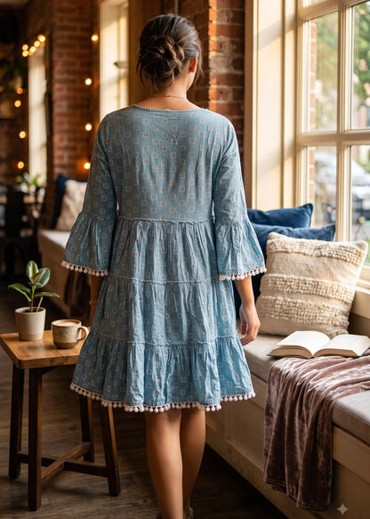 A woman viewed from behind wearing a light blue tiered dress with ruffled sleeves and hem, standing in a cozy sunlit room with brick walls, a window seat, pillows, and an open book.