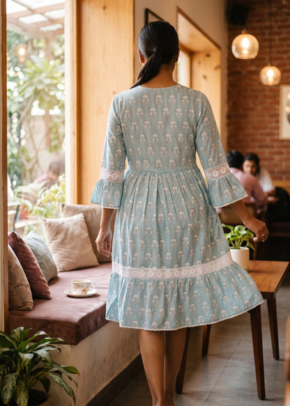 A woman viewed from behind wearing a light blue and white printed cotton dress with bell sleeves, standing in a cozy cafe interior with wooden windows, cushions, and pendant lights.