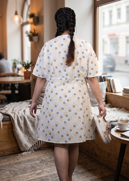 A woman viewed from behind wearing a white short kaftan sleeve dress with a blue and yellow floral pattern, walking inside a cozy, sunlit cafe with wooden floors and large arched windows.