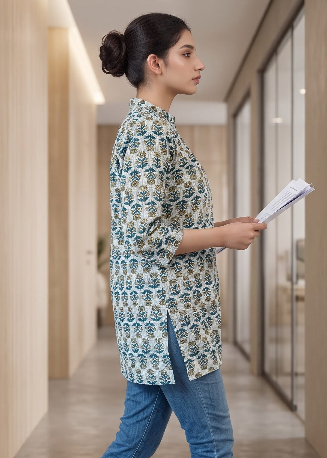 A woman in a patterned blue and beige short kurta, blue jeans, and white heels walks down a modern office hallway holding papers.