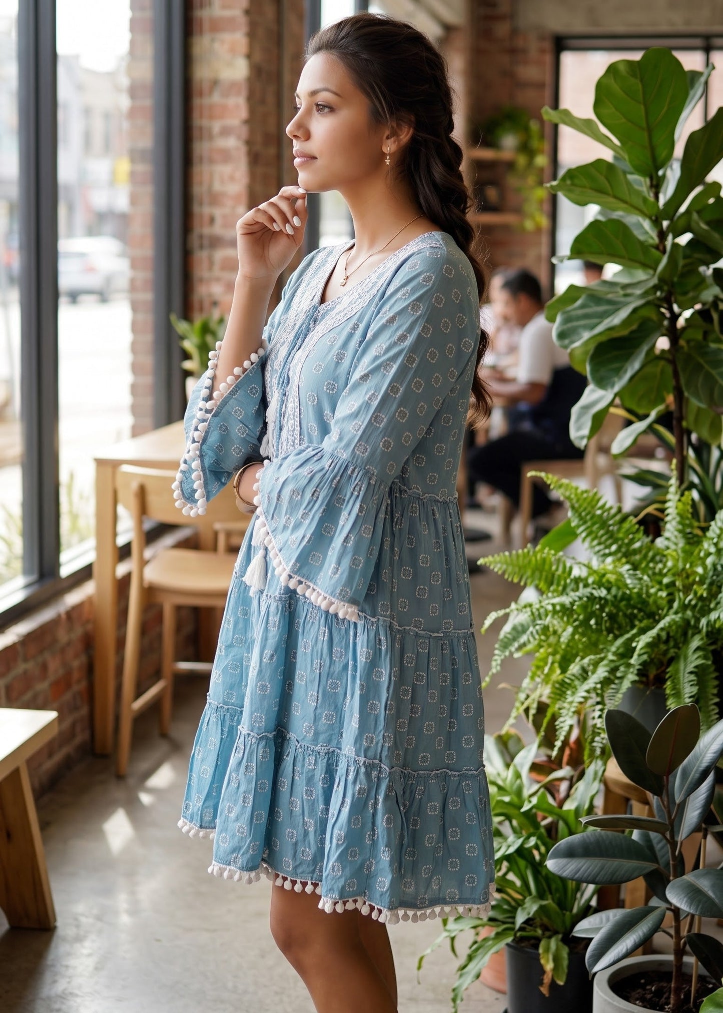 A woman wearing a light blue tiered bohemian-style dress with white floral embroidery and tassel trim, standing in a bright, modern loft cafe with brick walls and potted plants.