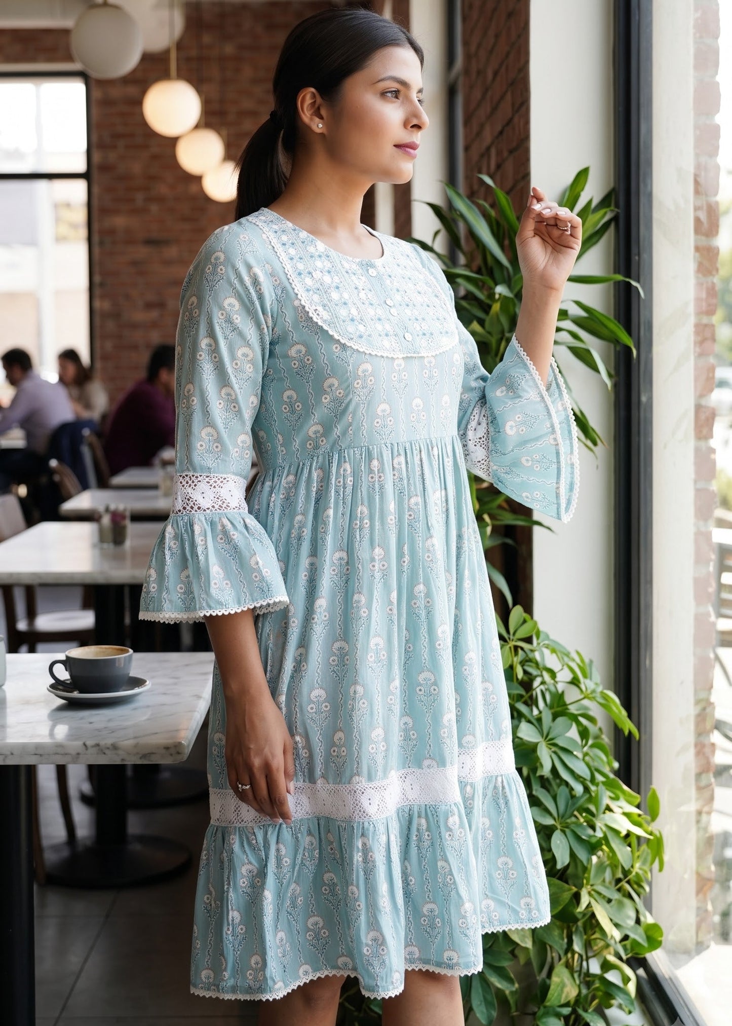 A woman wearing a light blue cotton short dress with white floral prints and lace trim, standing in a bright cafe next to a window.