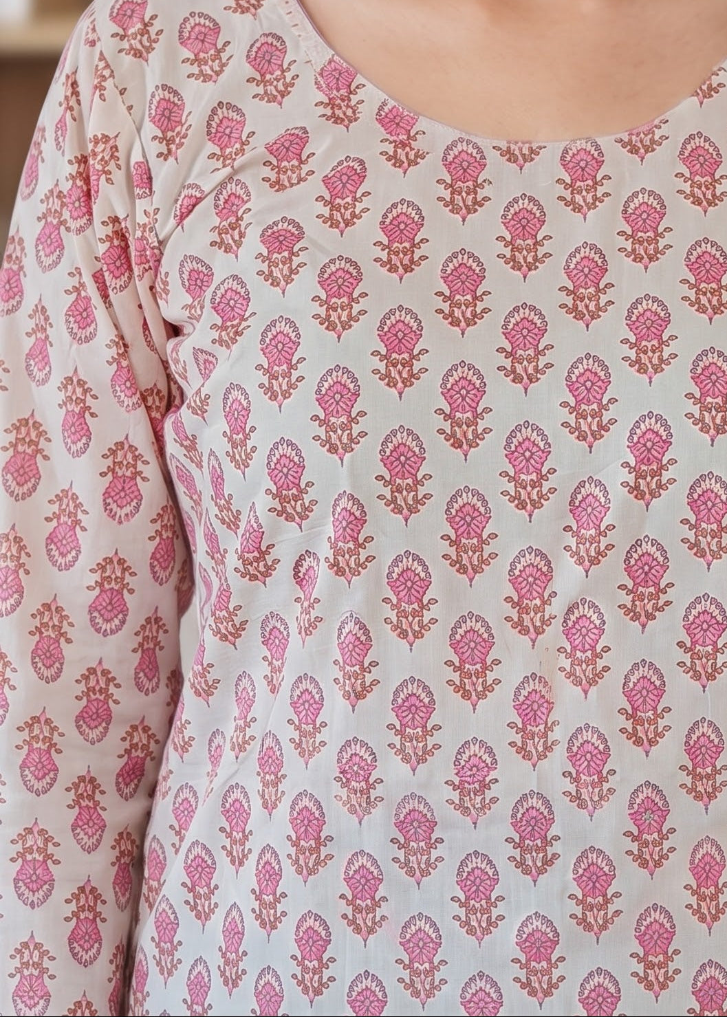 A woman with long dark hair wearing a white tunic short kurti top with pink floral block print pattern, photographed in a softly blurred indoor setting.