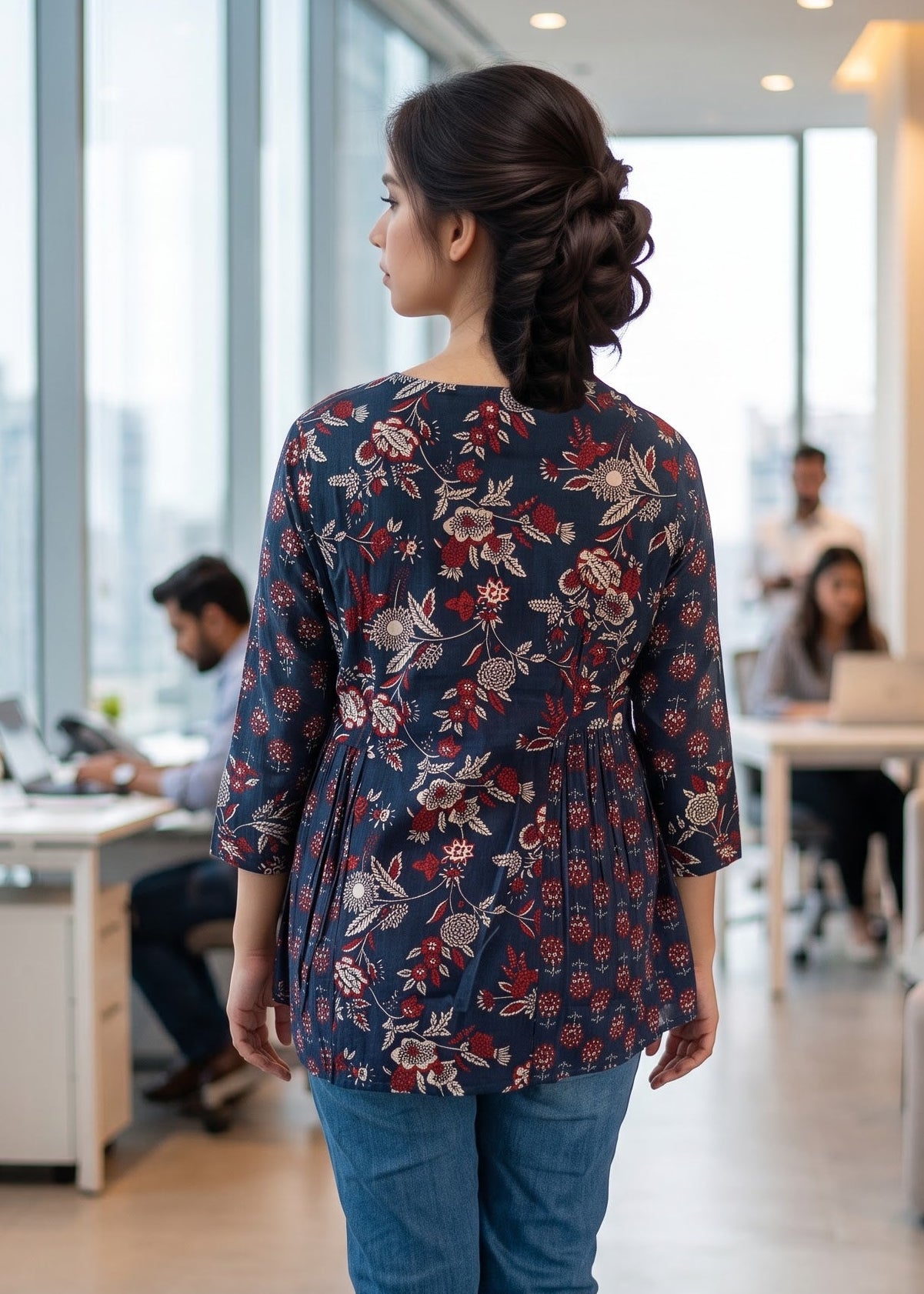 A woman viewed from behind wearing a navy blue floral three quater-sleeve short kurti tunic top and blue jeans, standing in a bright, modern office space with large windows and desks in the background.