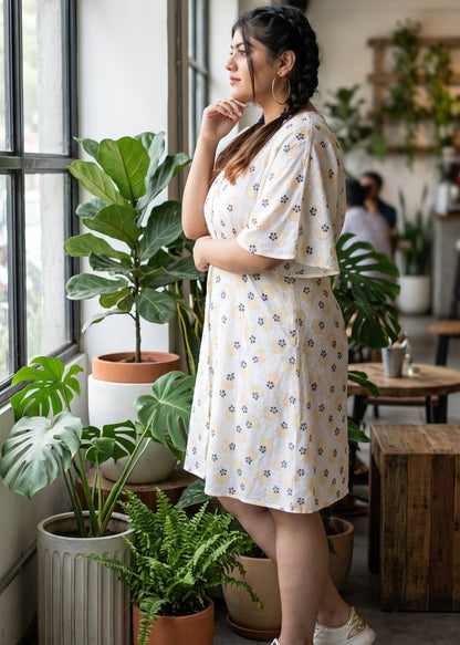 A woman stands by a large window in a bright indoor space, wearing a white colored short kaftan sleeve dress with a small blue and yellow floral & geometric pattern. She is surrounded by potted green plants in various pots, with wooden tables and a modern interior visible in the background.